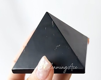 May include: A black obsidian pyramid with a polished, reflective surface. The pyramid is held in a hand against a white background. The pyramid's shape is a classic geometric form, with four triangular sides meeting at a point.