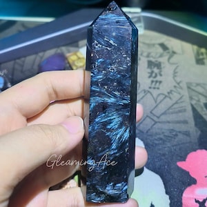 Könnte beinhalten: Ein dunkelblauer Kristallpunkt mit auffälligen blauen Adern. Der Kristall wird in einer Hand gehalten und zeigt seine polierte, geometrische Form. Der Text "Gleaming Ace" ist auf dem Kristall sichtbar. Dieser Kristall ist ein dekorativer Artikel.