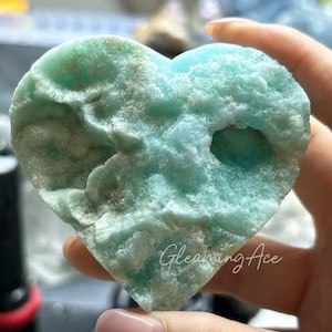 May include: A heart-shaped piece of blue-green celestite with a rough, textured surface and a small hole in the center. The stone is held in a hand, and the text "Gleaming Ace" is visible in the lower right corner.