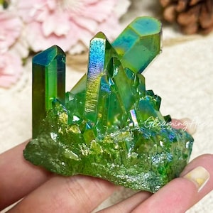 May include: A cluster of iridescent green aura quartz crystals with a metallic sheen. The mineral formation features multiple pointed vertical points rising from a textured base. The text GleamingAces is visible in white script on the crystal surface.