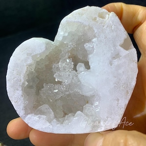 May include: A heart-shaped, white geode with a crystalline interior. The geode is held in a hand, showcasing its rough, textured exterior and the sparkling crystals within the cavity. The geode is a natural, decorative item.