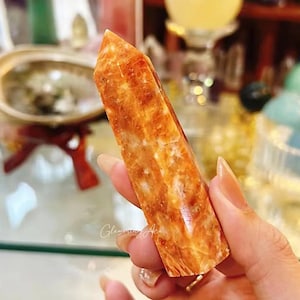 May include: A vibrant orange and white carnelian crystal point, with a smooth, polished finish. The crystal is held in a hand, showcasing its natural beauty and unique patterns.