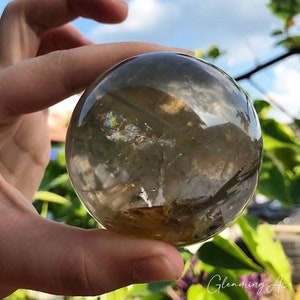 Può includere: Una sfera di cristallo di quarzo trasparente con una tonalità marrone fumè. La sfera è liscia e lucidata, e ha un leggero bagliore iridescente.