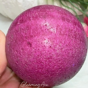 May include: A polished, spherical gemstone in shades of magenta and purple. The surface has a textured appearance. The text "Gleaming Ace" is visible on the bottom of the image. The background includes white and green foliage and pink flowers.