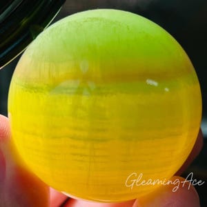 May include: A spherical object with a gradient of yellow and green. The object has a smooth surface and appears translucent. The text "Gleaming Ace" is written in cursive on the bottom right. The object is likely made of glass or a similar material.
