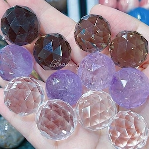 May include: A collection of faceted gemstone spheres in various shades of purple, brown, and clear. The spheres are arranged on a hand, showcasing their intricate facets and polished surfaces.