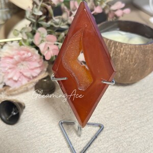 May include: A reddish-orange gemstone in a diamond shape, mounted on a silver metal stand. The gemstone has a geode-like centre with a lighter orange hue. The text "Gleaming Ace" is visible on the image.