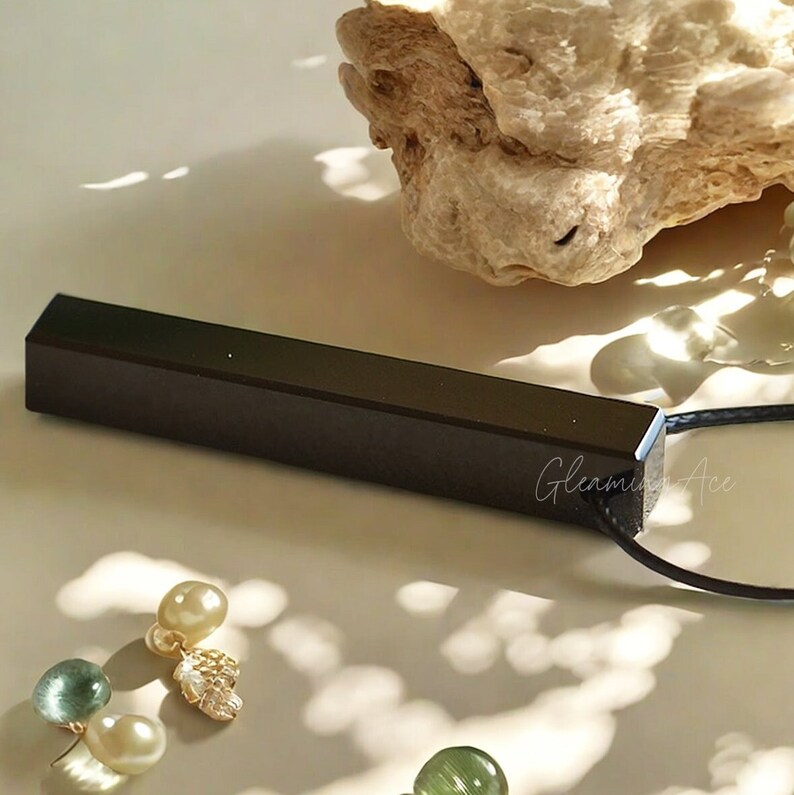 Puede incluir: Un colgante rectangular negro cuelga de un cord&oacute;n negro. El colgante est&aacute; hecho de madera y tiene un acabado liso y pulido. El colgante mide aproximadamente 5 cm de largo y 1,25 cm de ancho. El colgante est&aacute; colgando sobre una superficie blanca.