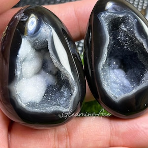 Peut inclure: Deux géodes en agate noire polie en forme de goutte d'eau. Chaque géode présente une cavité bordée de cristaux blancs et gris scintillants. Les géodes sont tenues dans une main, avec le texte "Gleaming Ace" visible.