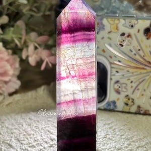 Könnte beinhalten: Ein hoher, sechseckiger Fluorit-Kristallturm mit horizontalen Bändern in Dunkelviolett, Magenta und durchscheinendem Weiß. Der Kristall hat eine polierte, glatte Oberfläche mit natürlichen inneren Rissen und dem Text Gleaming Ace.