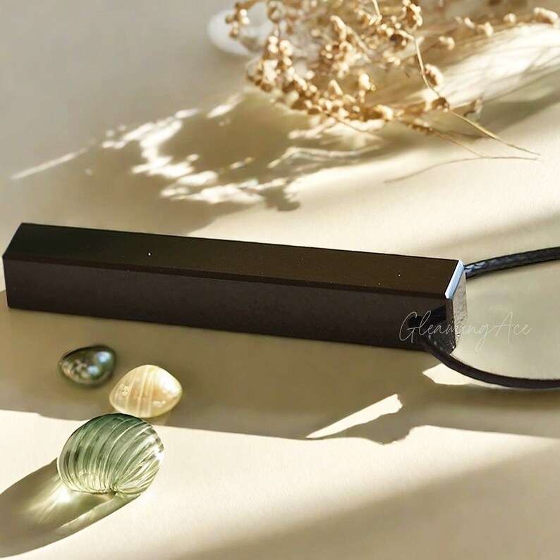 Puede incluir: Un colgante rectangular negro cuelga de un cord&oacute;n negro. El colgante est&aacute; hecho de madera pulida y tiene un acabado liso y brillante. El colgante descansa sobre una superficie blanca con peque&ntilde;as conchas cerca.