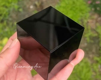May include: A solid black cube with a smooth, highly reflective surface. The object features sharp, defined edges and a glossy finish that mirrors its surroundings. The script text "Gleaming Ace" is printed in white on the front face.