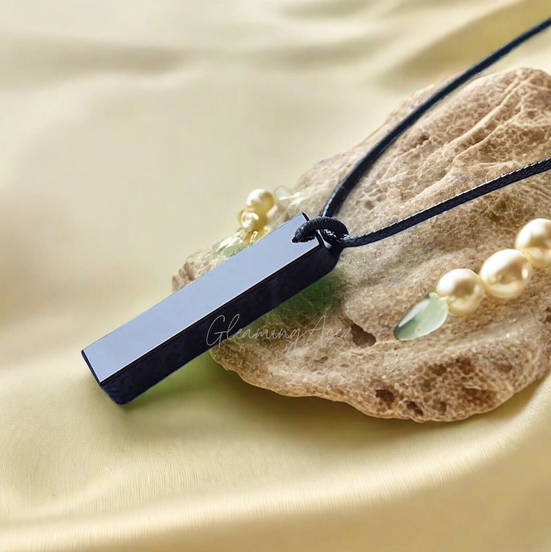 Puede incluir: Un colgante de barra de obsidiana negra en un collar de cord&oacute;n negro. El colgante descansa sobre un trozo de coral con piedras blancas y verdes.