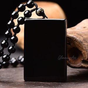Peut inclure: Un collier avec un pendentif en obsidienne noire de forme carrée et des perles noires. Le pendentif porte des inscriptions dorées.