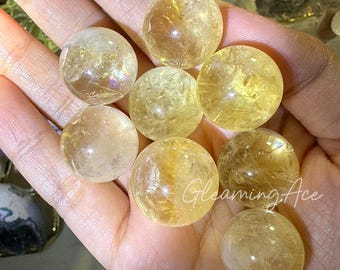 May include: A collection of eight yellow citrine crystal spheres, ranging in size, displayed on a hand. The spheres are smooth and translucent, with a slight golden hue.