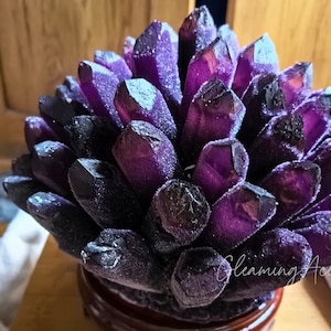 Könnte beinhalten: Ein Cluster aus tiefvioletten Amethystkristallen, die in einer kreisförmigen Form angeordnet sind und einer Blume ähneln. Die Kristalle sind auf einem kleinen, dunkelbraunen Holzsockel befestigt.