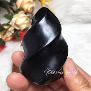 Pode incluir: Uma escultura de pedra preta polida com um design suave e rodopiante. A escultura tem um acabamento brilhante e é segurada em uma mão. O texto "Gleaming Ace" está escrito no canto inferior direito da imagem.