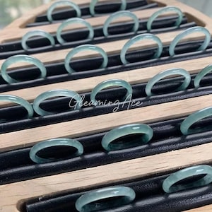 May include: A display of light green jade rings arranged on a wooden tray with black slots. The rings are smooth and translucent, with a subtle glow. The text "Gleaming Ace" is visible on the tray.