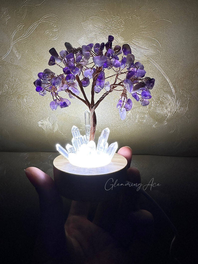 May include: A purple amethyst crystal tree with a white light base and clear quartz crystals. The tree is on a wooden base and is lit up. The text "Gleaming Ace" is visible in the image.