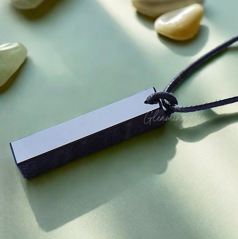 Puede incluir: Un colgante rectangular negro con una superficie lisa y pulida cuelga de un collar de cord&oacute;n negro. El colgante est&aacute; sobre una superficie verde claro.