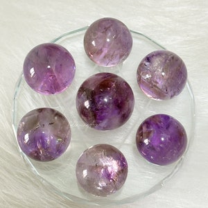May include: Seven amethyst spheres of varying shades of purple and white, arranged on a clear glass plate.