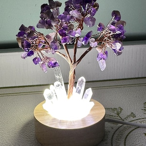 May include: A purple amethyst crystal tree with a copper wire base and a white crystal base. The tree is lit from below with a white light. The tree is sitting on a wooden base.