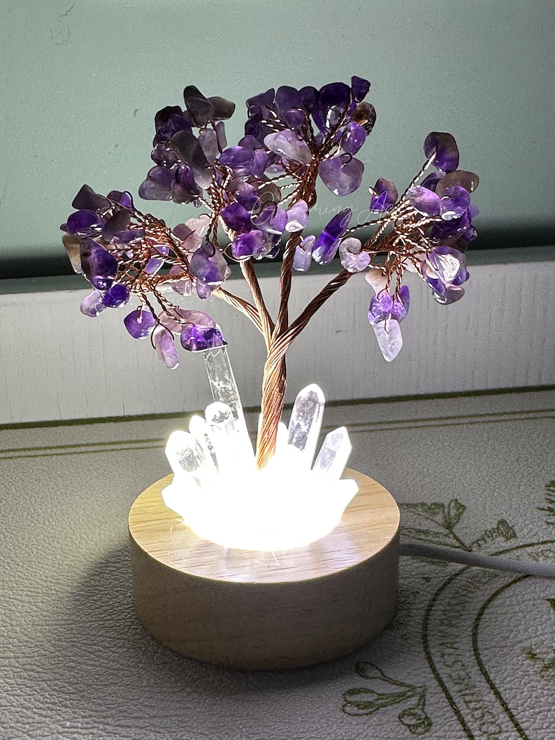 May include: A purple amethyst crystal tree with a copper wire base and a white crystal base. The tree is lit from below with a white light. The tree is sitting on a wooden base.
