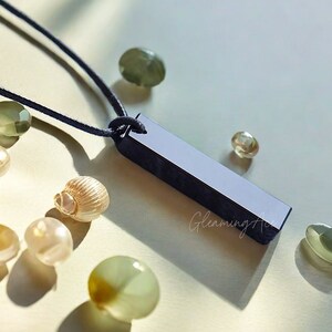 Puede incluir: Un collar con un colgante de barra negra en un cord&oacute;n negro. El colgante es largo y rectangular, con una superficie lisa y pulida.