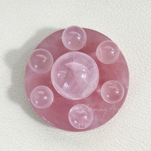 May include: A pink rose quartz crystal sphere sits on a pink rose quartz base with six smaller rose quartz spheres surrounding it.
