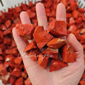 Puede incluir: Primer plano de una mano sosteniendo piedras de jaspe rojo en bruto. Las piedras tienen una apariencia natural sin pulir. El texto "Gleaming Ace" es visible en la imagen.