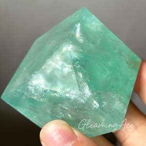 Könnte beinhalten: Ein grüner Fluorit-Kristallwürfel mit schillernden Facetten. Der Kristall ist glatt und poliert, in einer hellgrünen Farbe.