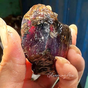 May include: A raw, uncut gemstone with a dark, translucent appearance. The stone has a mix of deep purple, red, and brown hues, with a glossy, wet-like sheen. The surface is uneven and textured, revealing the natural formation of the mineral.