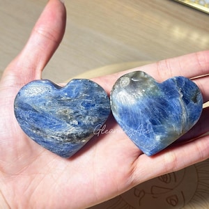 Puede incluir: Dos piedras de cianita pulidas en forma de corazón con vetas de color azul oscuro y gris y un acabado liso y brillante. El texto Gleaming Ace es visible en letra blanca sobre el centro de las dos piedras, que muestran texturas minerales naturales.