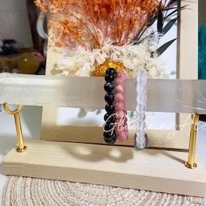 May include: A wooden display stand with gold supports holds three beaded bracelets. The bracelets are black, pink, and clear. The background includes dried flowers and a framed picture.