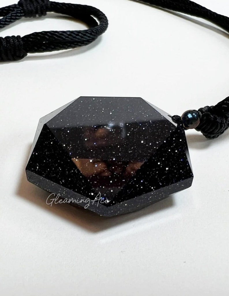 May include: A black gemstone pendant with a faceted, geometric shape. The pendant is made of black stone with a glittery finish and is suspended from a black cord.