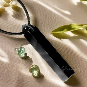 Puede incluir: Collar con colgante de obsidiana negra de forma rectangular y cord&oacute;n negro. El colgante est&aacute; pulido y tiene una superficie lisa. El collar se muestra sobre una superficie marr&oacute;n claro con piedras verdes.