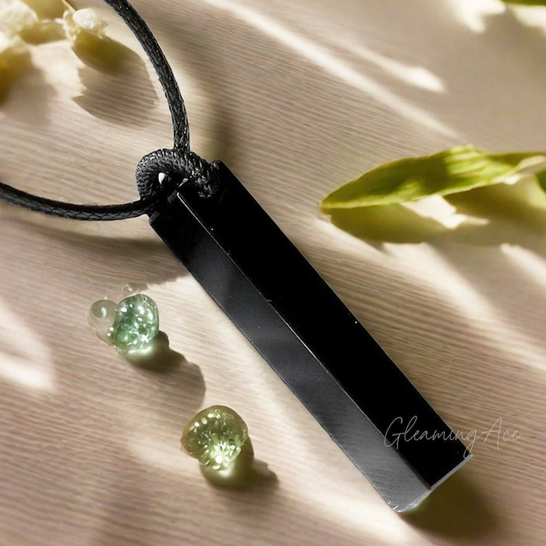 Puede incluir: Collar con colgante de obsidiana negra de forma rectangular y cord&oacute;n negro. El colgante est&aacute; pulido y tiene una superficie lisa. El collar se muestra sobre una superficie marr&oacute;n claro con piedras verdes.