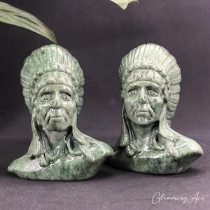 May include: Two green jade stone carvings of Native American chiefs wearing headdresses. The carvings are detailed and realistic, with expressive faces and intricate headdresses.