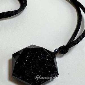 May include: A black, seven-sided pendant with a sparkly, starry texture hangs from a black cord. The pendant is shaped like a heptagon.