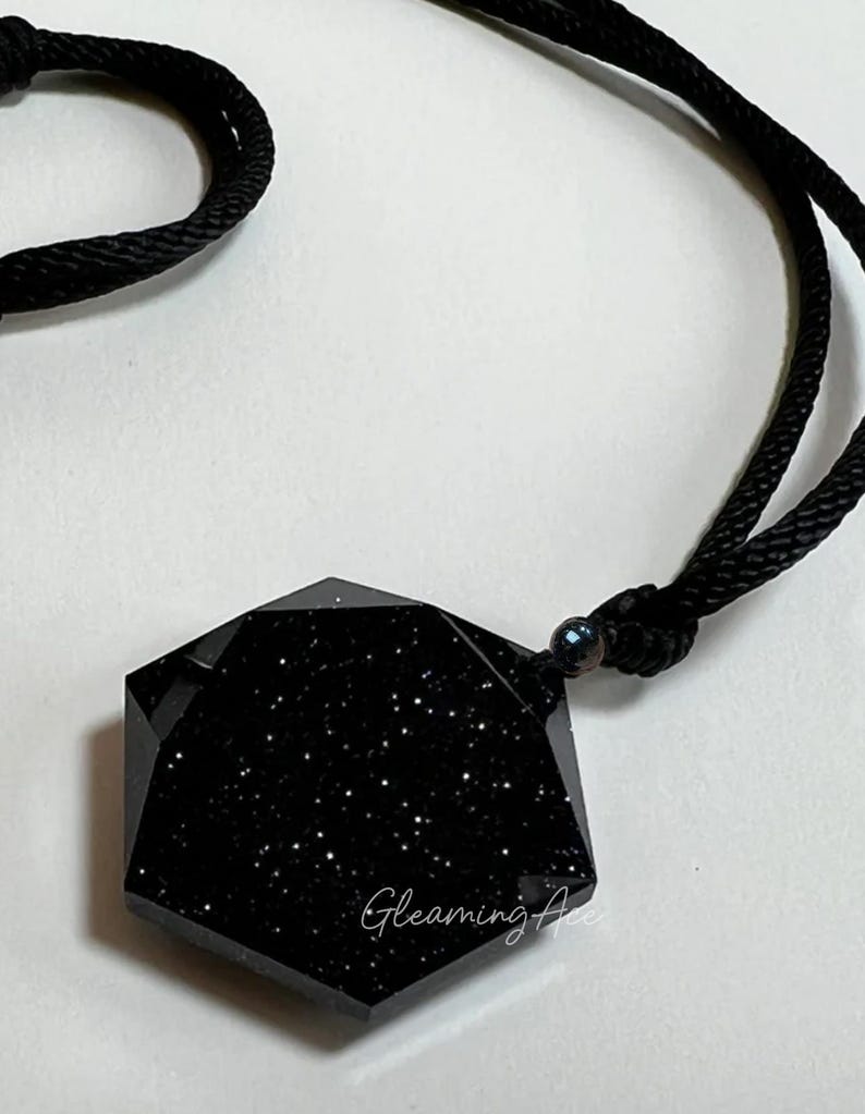 May include: A black, seven-sided pendant with a sparkly, starry texture hangs from a black cord. The pendant is shaped like a heptagon.
