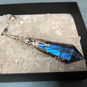 Pode incluir: Um pêndulo de cristal azul e prateado com um design metálico detalhado. O pêndulo está pendurado em uma corrente prateada e está descansando em um leito de cristais brancos.