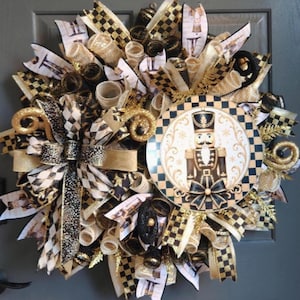 May include: A decorative wreath featuring a central circular sign with a nutcracker illustration, surrounded by layers of black, gold, and white mesh ribbons. The design includes patterns of black and white checks and nutcracker motifs.