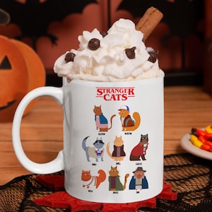 May include: White ceramic mug with the text "Stranger Cats" and illustrations of cats dressed as characters. The mug is topped with whipped cream, chocolate chips, and a cinnamon stick. The mug is on a red coaster.