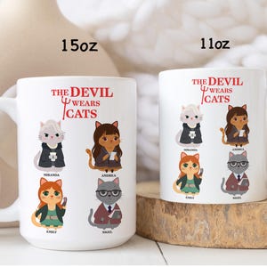 May include: Two white ceramic mugs with the text "THE DEVIL WEARS CATS" and illustrations of cats dressed in business attire. One mug is labeled 15oz, the other 11oz. The cats are named Miranda, Andrea, Emily, and Nigel.