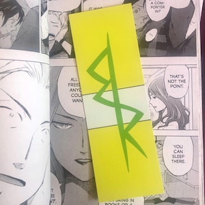 May include: A bright yellow bookmark with a green geometric design and a white stripe. The bookmark is placed on top of a manga comic book. The comic book pages are open, revealing black and white illustrations and speech bubbles.