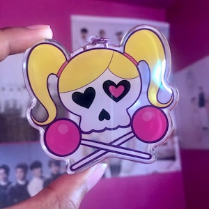 May include: A clear acrylic keychain featuring a cartoon skull with pigtails, hearts for eyes, and two pink lollipops crossed behind the skull.
