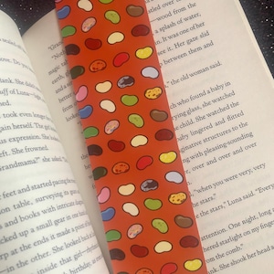 May include: A red bookmark with a repeating pattern of colorful jelly beans. The bookmark is approximately 10.5 cm wide and 15 cm tall.