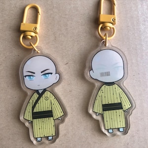 以下が含まれることがあります： 2つの漫画のキャラクターのキーホルダー。各キーホルダーは、黄色の着物スタイルの衣装を着たハゲのキャラクターが特徴です。キーホルダーには金色の留め金が付いています。1つのキャラクターは青い目を持ち、もう1つは顔にバーコードのようなデザインがあります。