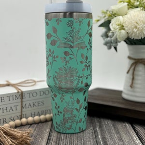 May include: A teal stainless steel tumbler with a lid and a straw. The tumbler has a design of books and flowers in a silver color.