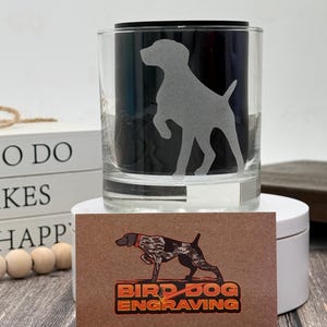 May include: A clear glass tumbler with a frosted silhouette of a dog. The tumbler has a black interior. A business card with a dog illustration and the text "BIRD DOG ENGRAVING" is in front of the tumbler.